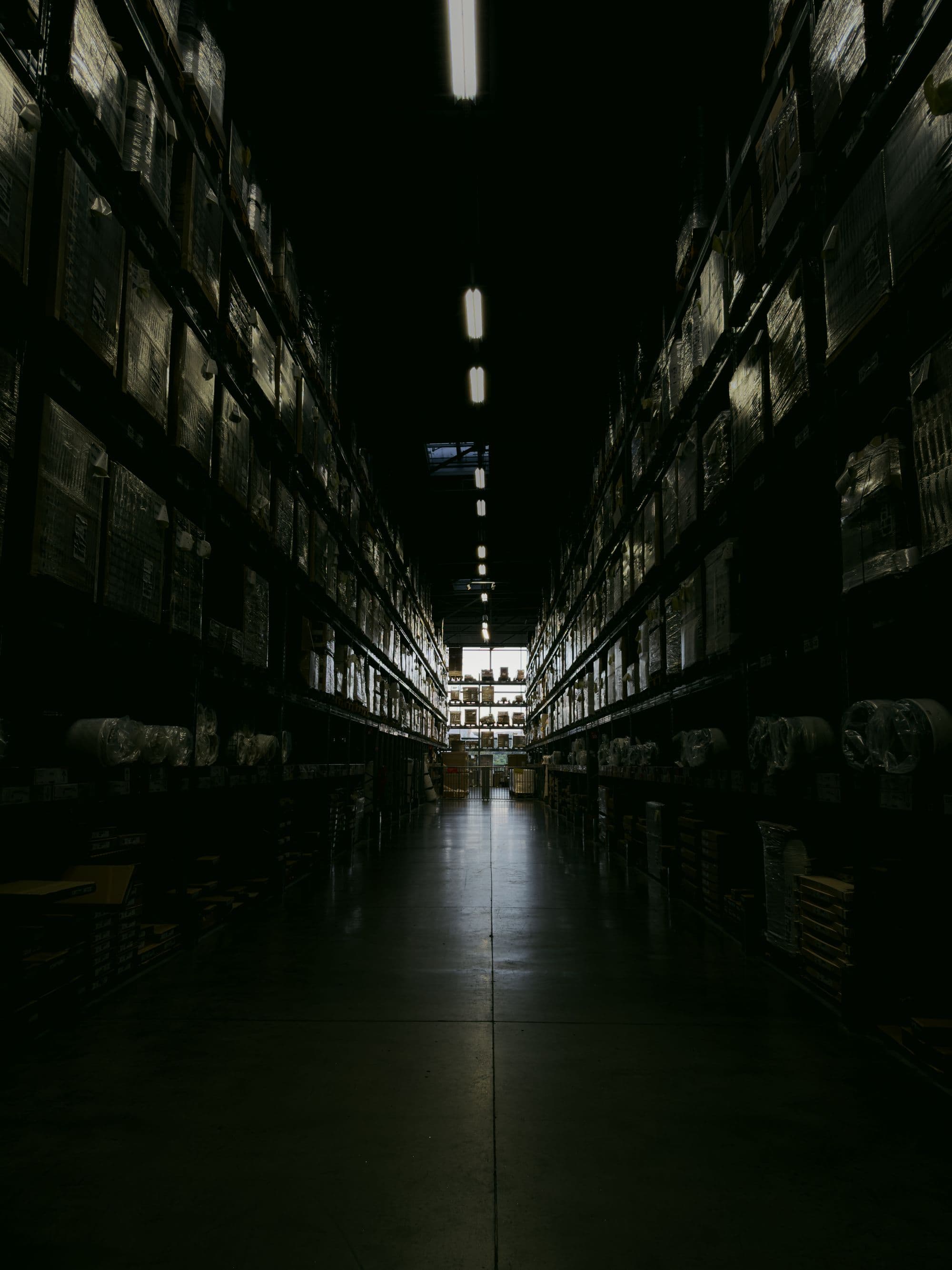 A dimly lit warehouse aisle between tall loaded racks, a line of ceiling lights running away into the depth.