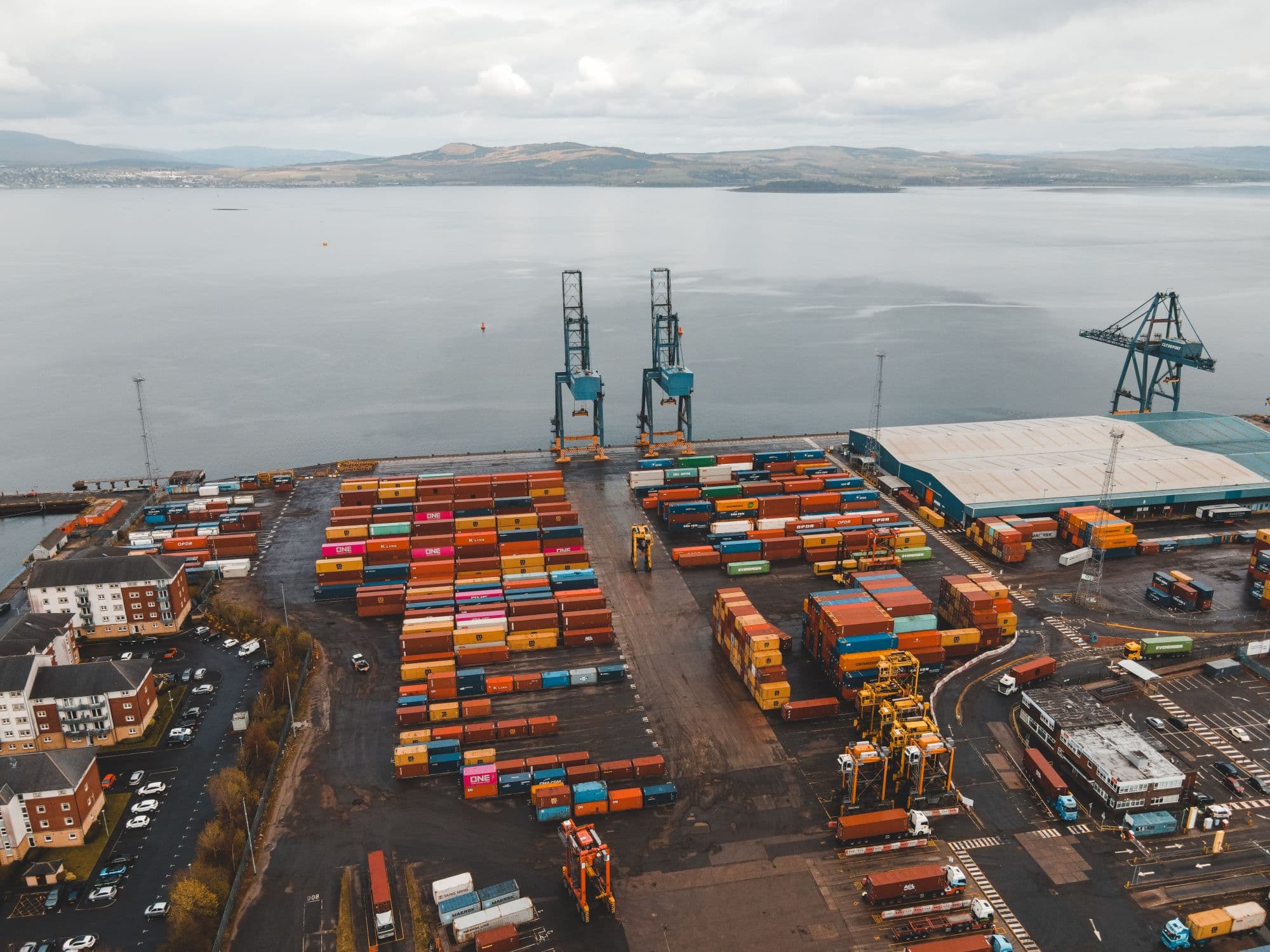 Aerial view of a container terminal on an estuary: rows of coloured containers, two quay cranes and a shed, hills on the horizon.
