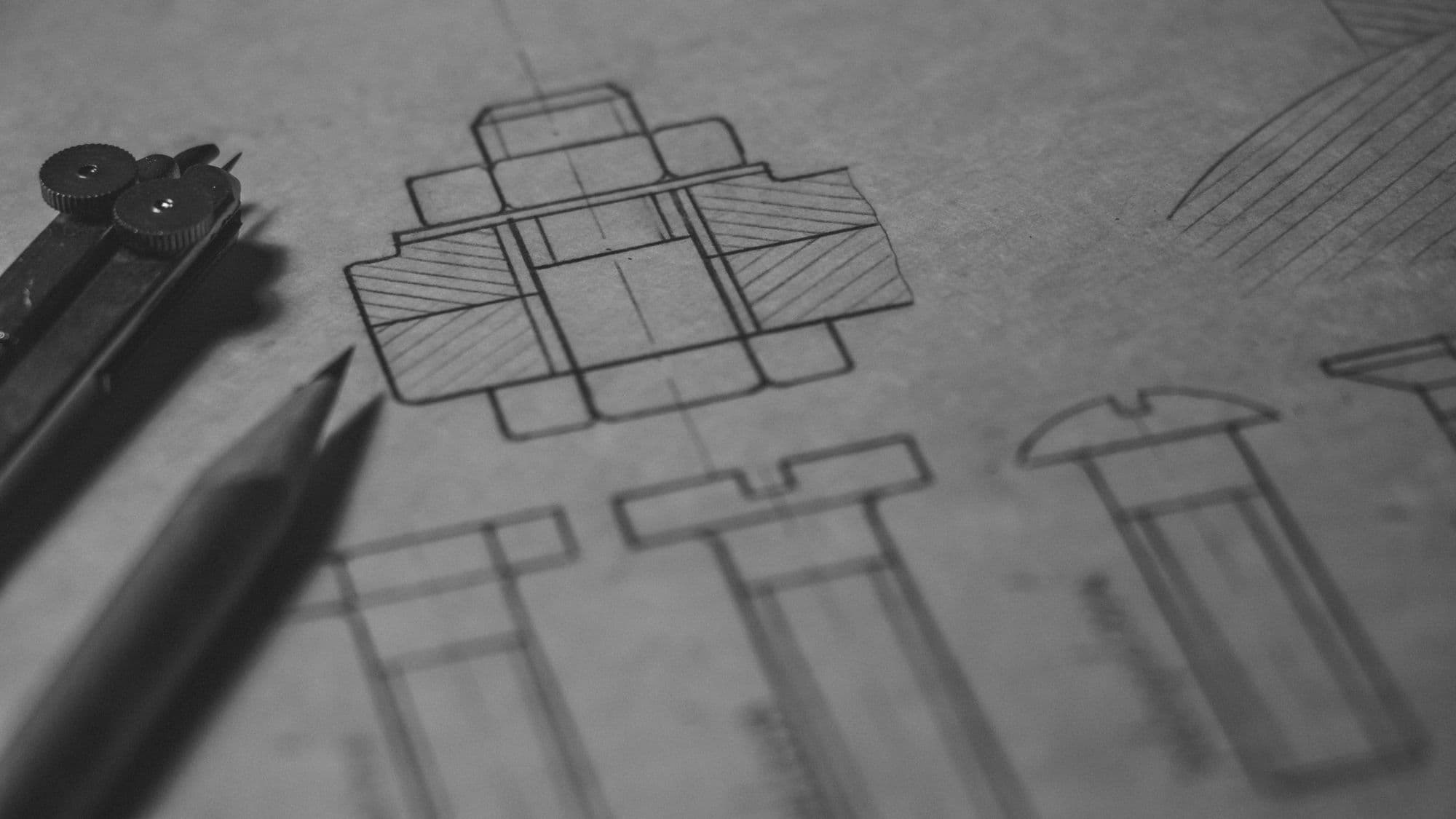 A pencil engineering drawing of machined parts and bolts in multiple views, with a compass and two pencils resting on grey paper.