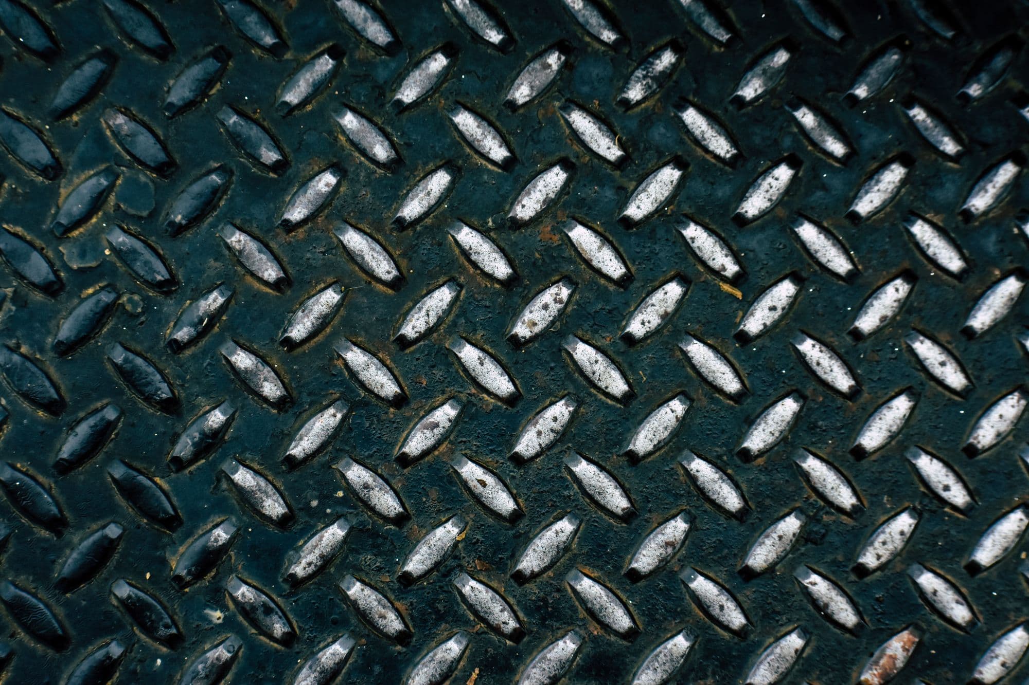 Close view of a dark checker-plate steel sheet, its diamond ribs catching light against a near-black ground.
